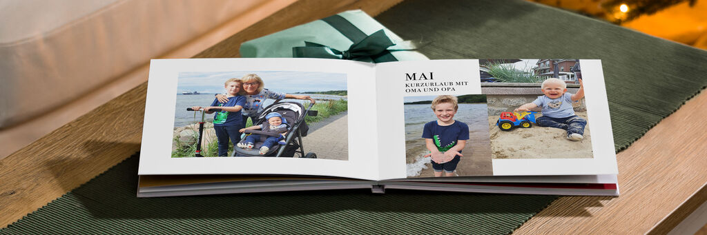 Ein geöffnetes Fotobuch mit zwei Bildern von Kindern, die draußen spielen. Das Fotobuch liegt auf einem Tisch, umgeben von einer grünen Decke und einem Geschenk mit einem grünen Band. Die Kinder freuen sich und zeigen aktiv ihr Spielzeug.