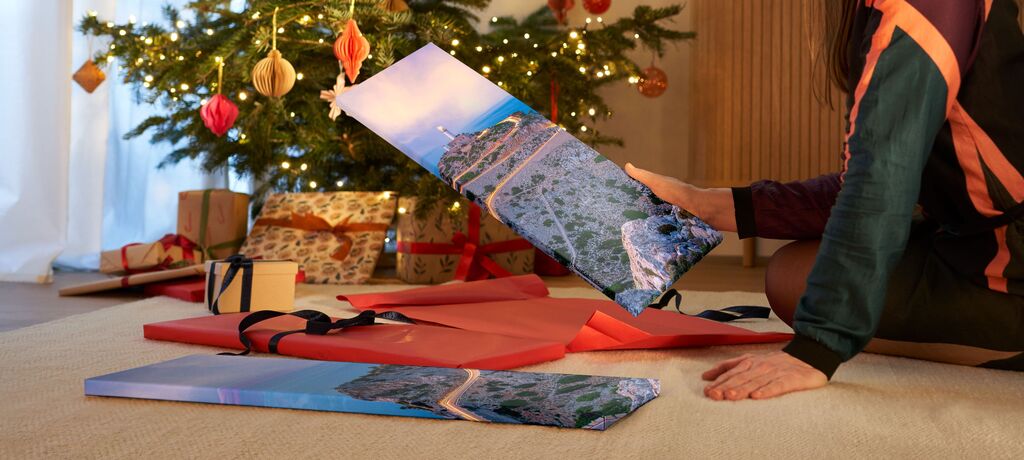 Person hält ein Geschenk mit einem Bild von einer Küstenlandschaft vor einem festlich dekorierten Weihnachtsbaum