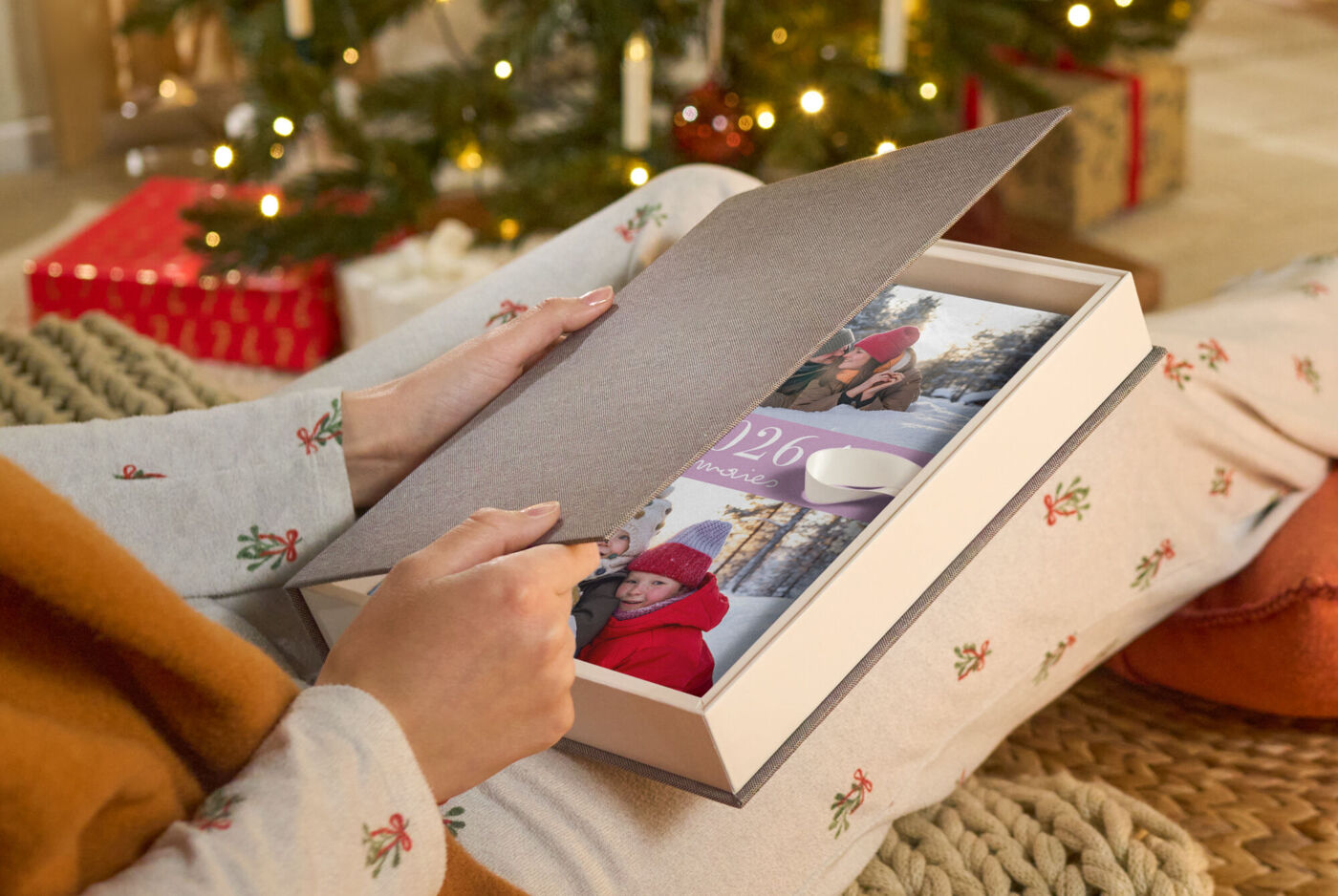 Hände öffnen eine hochwertige Geschenkbox mit einem CEWE FOTOUCH auf einem Holztisch, im Hintergrund ein festlich geschmückter Baum
