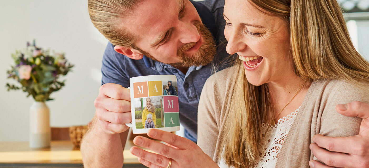 Liebende Person gibt Tasse mit Namen "MAMA" an eine andere Person, die lächelt