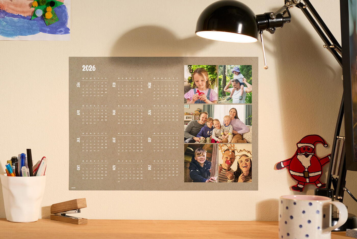 Echte Alltagswärme am Schreibtisch: persönlicher Jahreskalender mit Familienfotos, warmes Lampenlicht, Tasse und kleinem Weihnachtsmann.