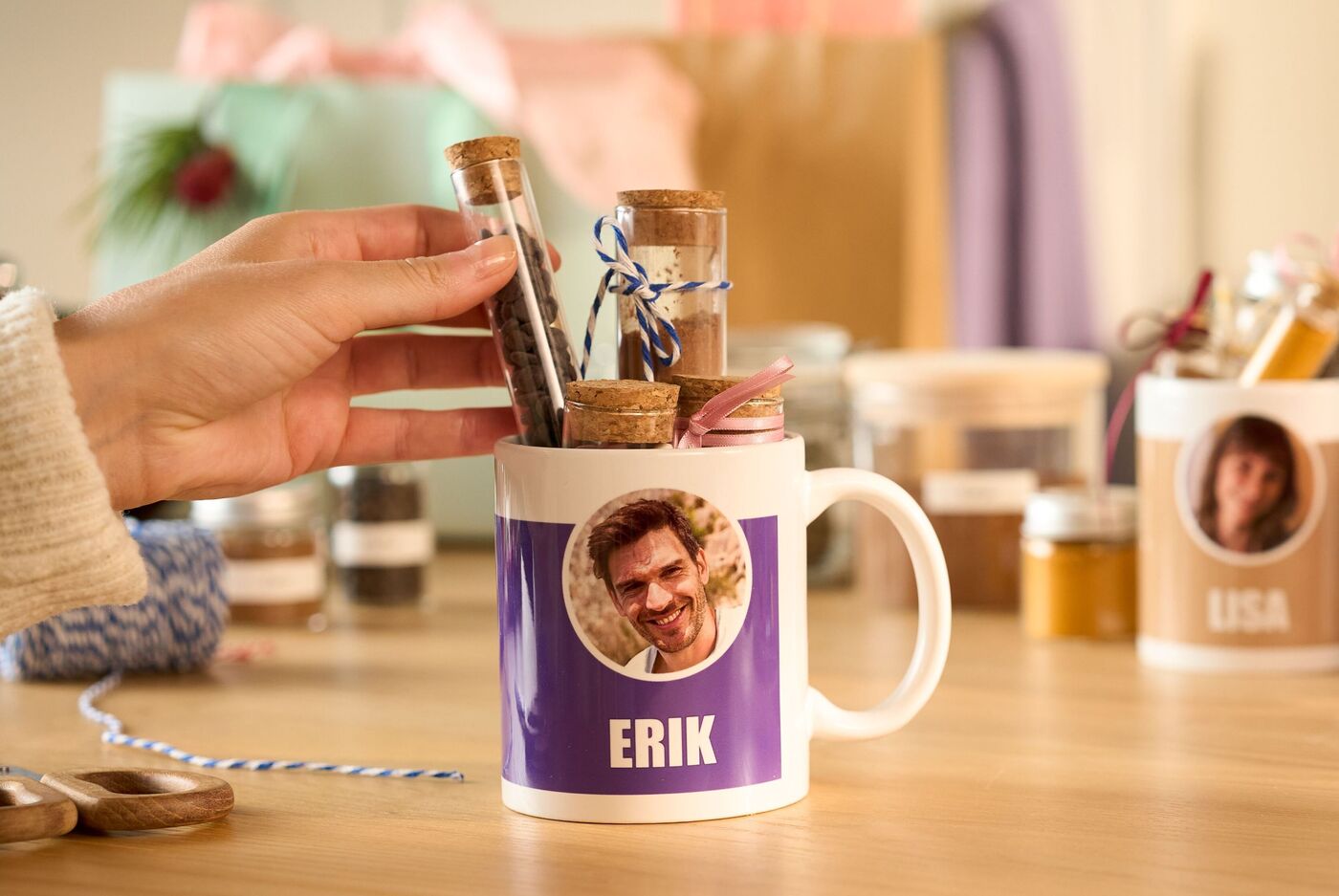 Eine lila und weiße Tasse mit Foto eines Mannes und dem Namen "Erik" steht auf einem Tisch, eine Hand befüllt sie mit kleinen Geschenken