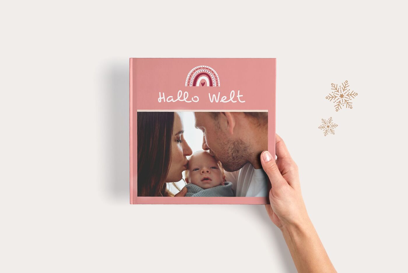 Hand hält ein rosa CEWE FOTOBUCH Quadratisch zum Thema Baby, mit Regenbogenmotiv, Fotofenster und Titel "Hallo Welt"