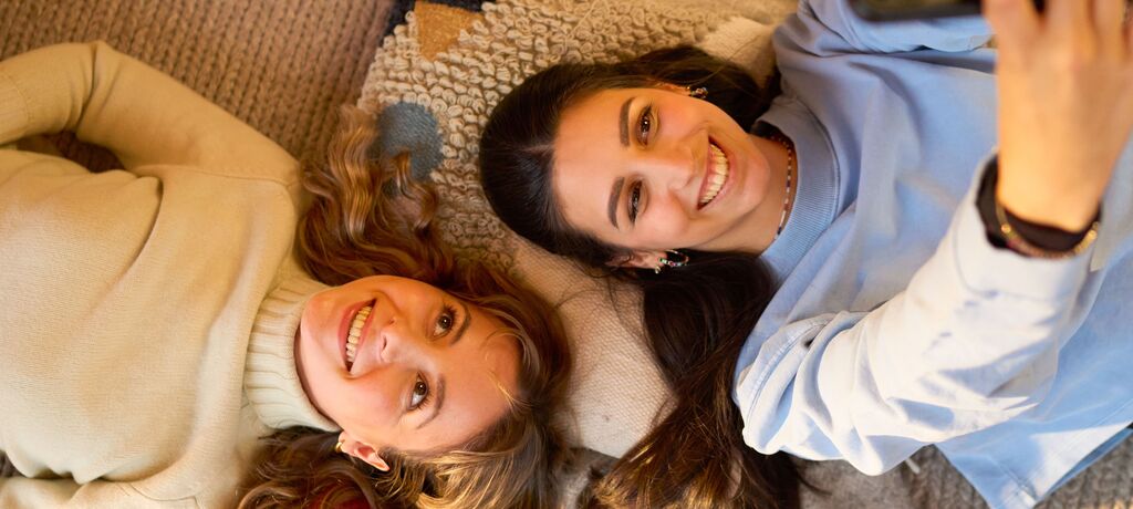 Zwei fröhliche Frauen liegen auf einem gemütlichen Teppich und machen ein Selfie. Ihre Freude strahlt durch das Bild.