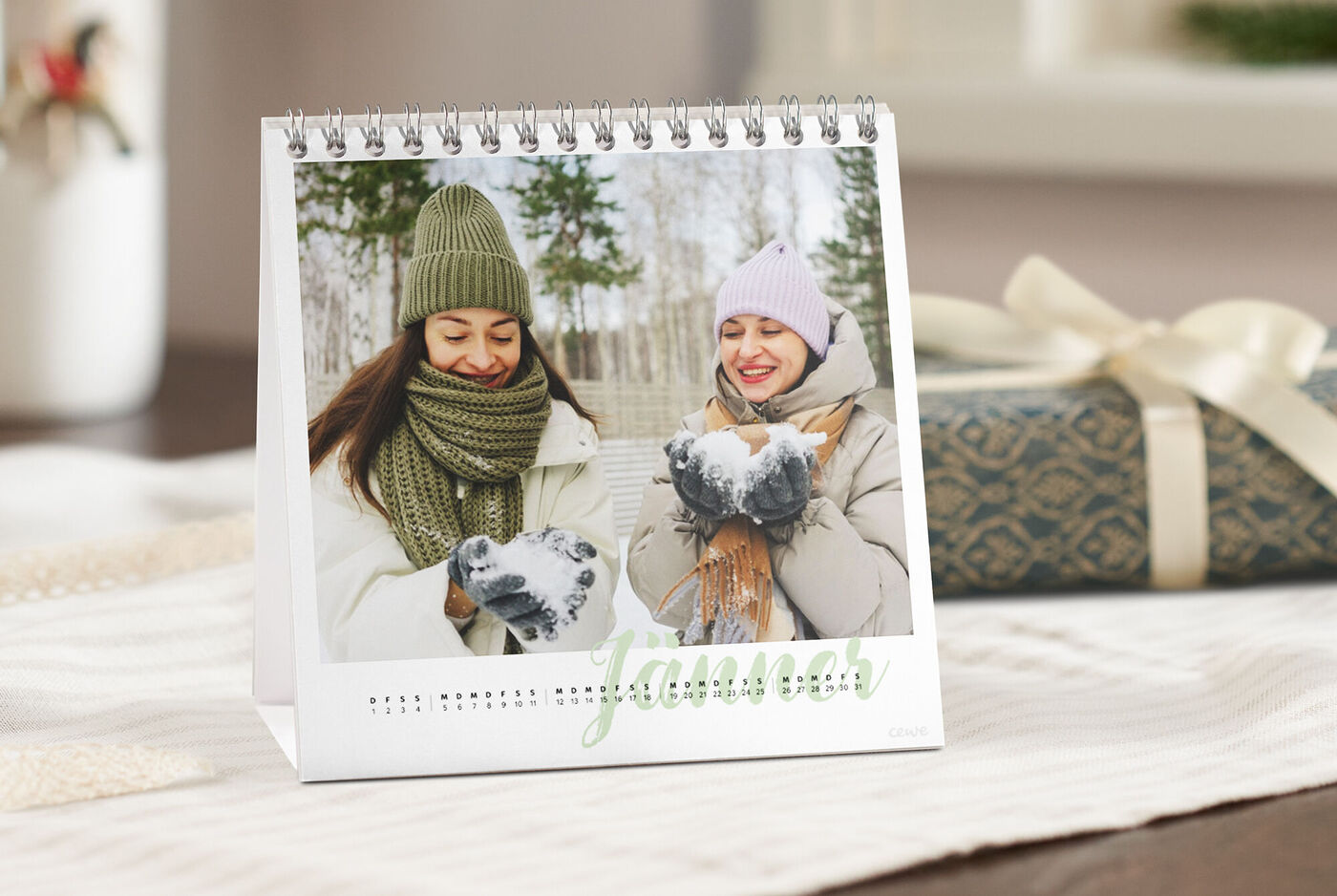 CEWE Tischkalender mit Jänner-Motiv: Zwei Personen in winterlicher Kleidung, Schneeball in den Händen, daneben ein Geschenk