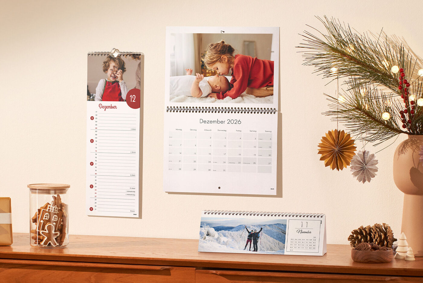 Drei CEWE Kalender unterschiedlicher Formate an einer Wand und auf einem Regal mit Weihnachtsdekoration, Plätzchen und Tannenzweigen
