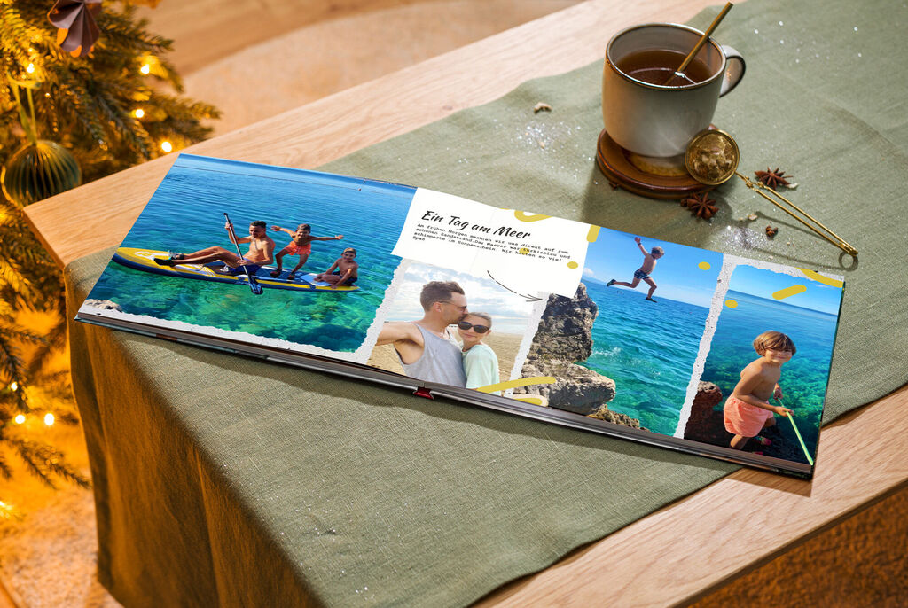 Ein CEWE FOTOBUCH auf einem Tisch, mit Bildern von einem Ausflug am Wasser. Eine Tasse steht daneben.