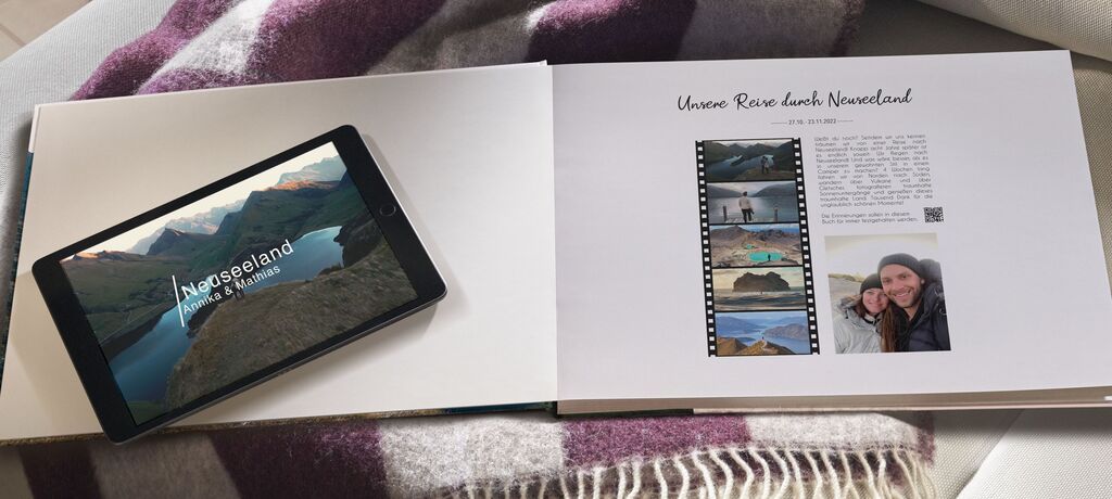 Ein aufgeschlagenes Fotobuch liegt auf einer Wolldecke. Ein darauf liegendes Tablet zeigt ein Neuseeland-Motiv.