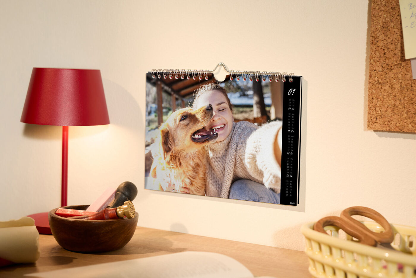 Wandkalender über einem gemütlichen Schreibtisch; Foto zeigt eine Person im Strickpulli, die ihren Hund liebevoll an sich drückt – warmer, r