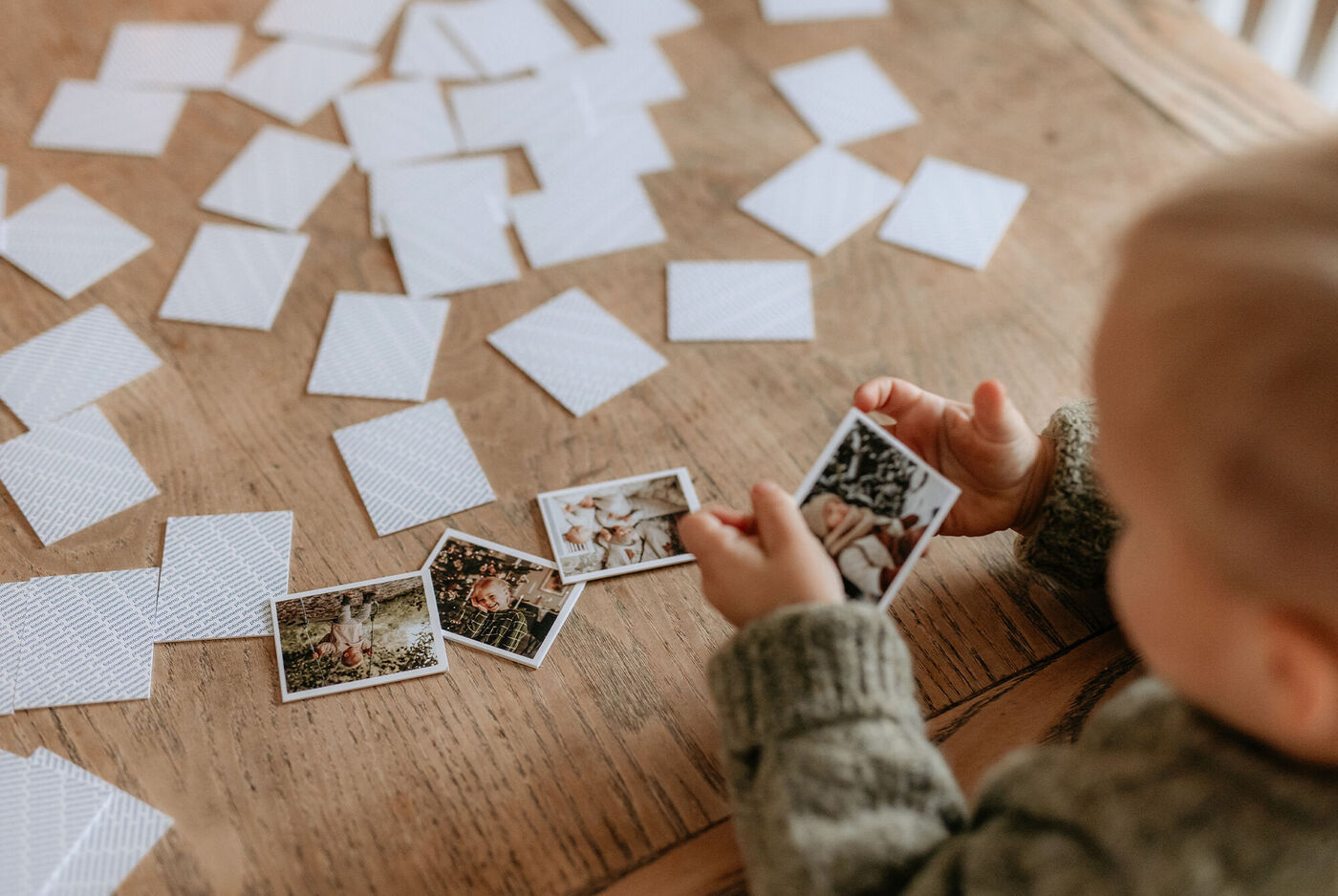 Ein Kind hält zwei kleine Fotos in Händen, während mehrere leere Fotokarten auf einem Tisch liegen.