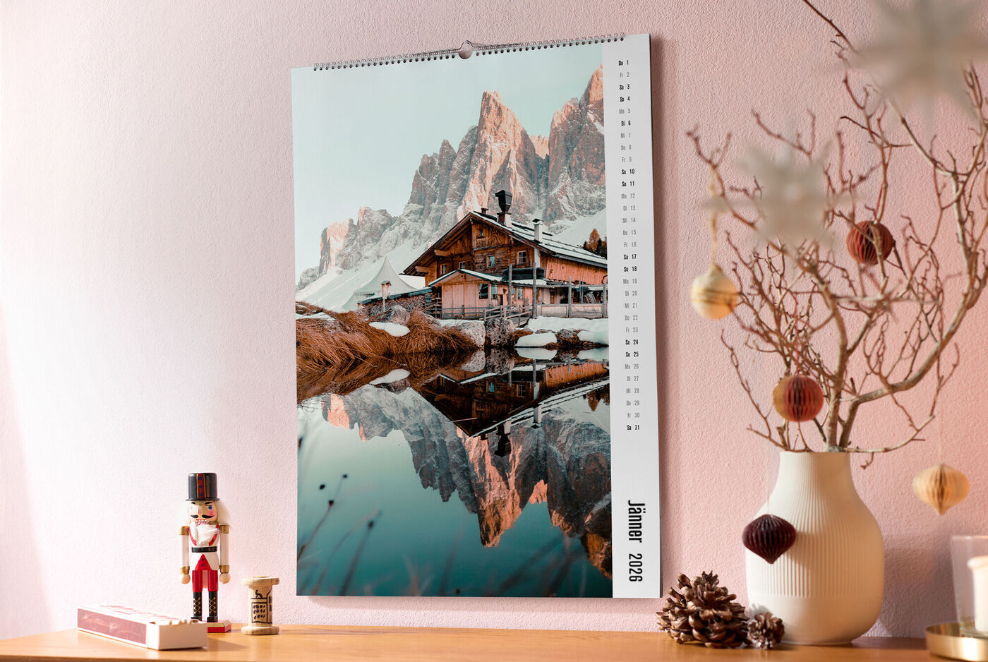 CEWE Fotokalender mit Berghütte und Spiegelung im See, daneben Nussknacker, Zapfen und Vase