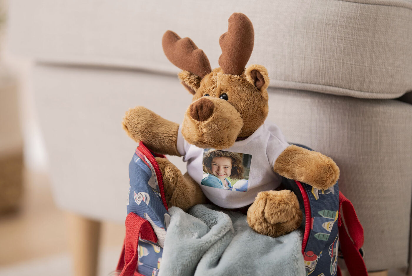 Ein brauner Teddybär mit Geweih sitzt auf einer Tasche, umgeben von einem weichen grauen Tuch. Der Teddybär trägt ein T-Shirt mit einem Bild.