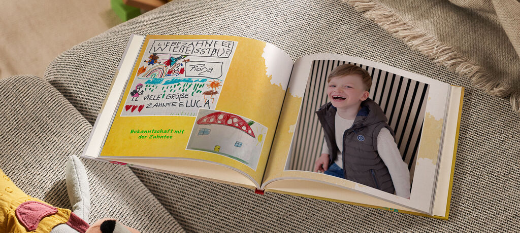 Ein geöffnetes CEWE Fotobuch mit bunten Zeichnungen und einem lächelnden Kind in grauer Weste.
