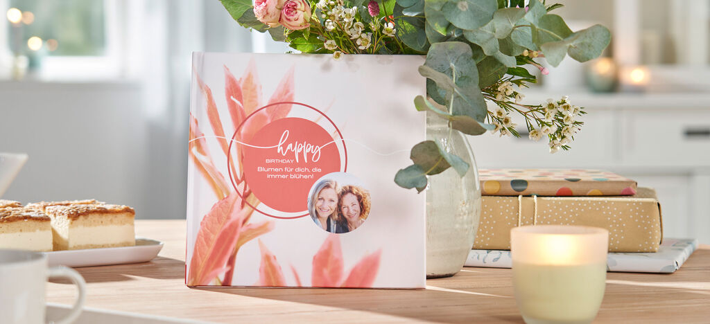 Ein CEWE Fotobuch auf einem Holztisch, umgeben von Blumen, Kerzen und Leckereien, strahlt Freude und Wertschätzung aus