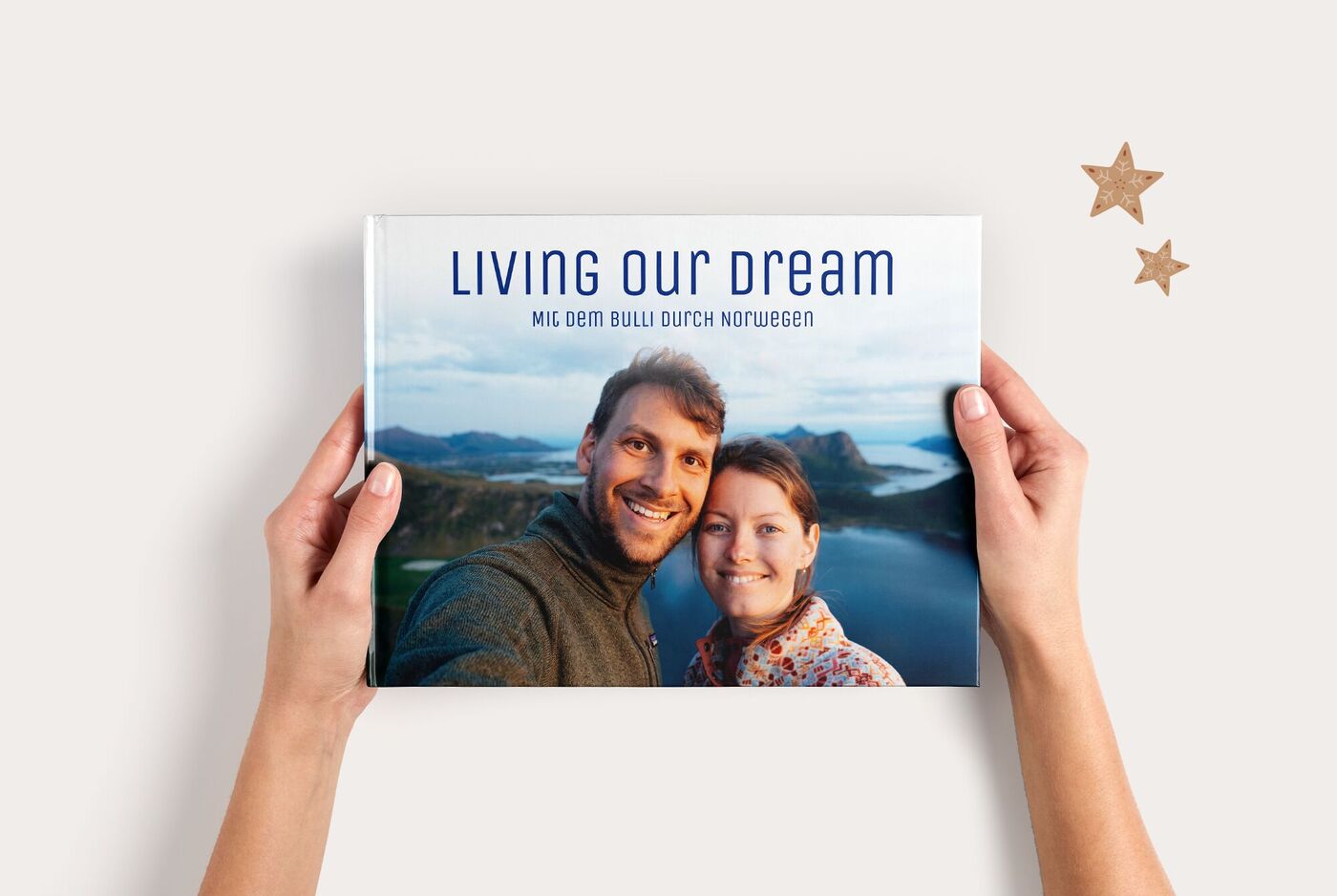 Zwei Hände halten ein CEWE FOTOBUCH Groß Panorama mit einem Paarfoto vor einem Fjord und dem Titel "LIVING OUR DREAM"