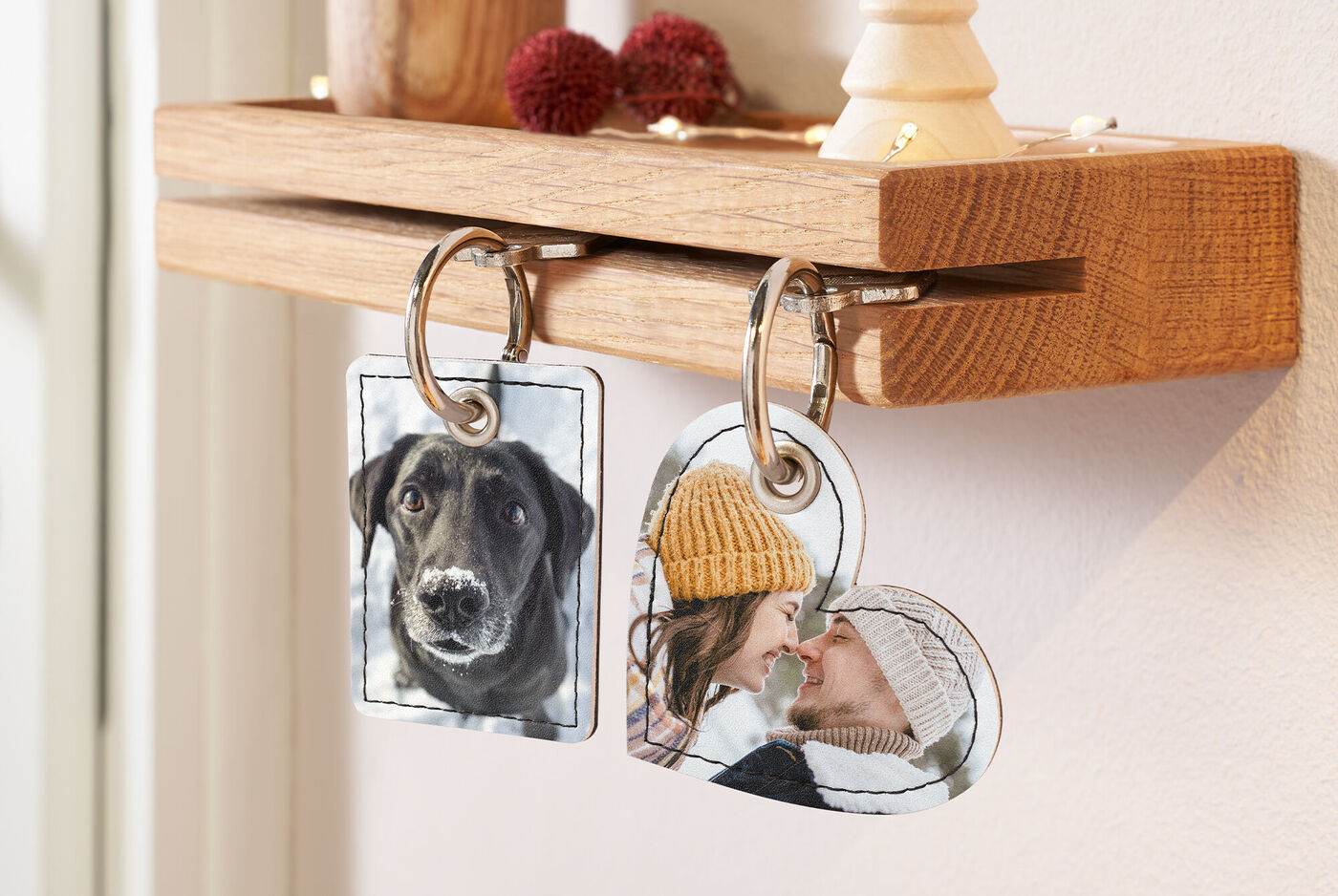 Zwei individuelle Schlüsselanhänger hängen an einem Holzregal. Der linke Anhänger zeigt einen Hund, der rechte Anhänger zeigt eine Person mit Mütze. Beide Anhänger sind in Form eines Rechtecks und eines Herzens.