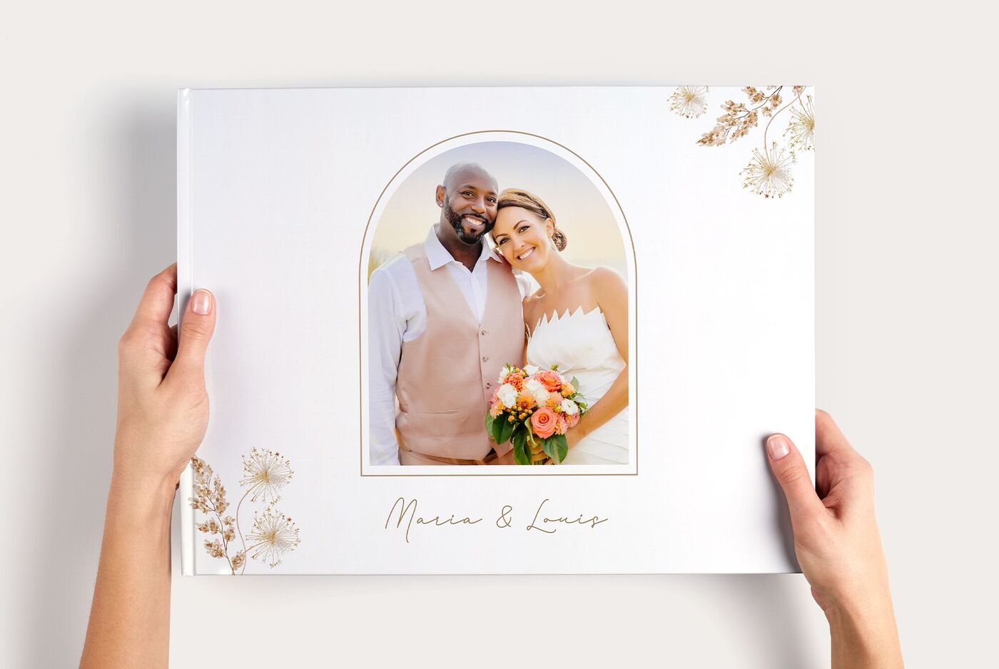 Zwei Hände halten Hochzeits- CEWE FOTOBUCH XXL Panorama mit Paarfoto in Bogenrahmen und Schriftzug "Maren & Lucas"