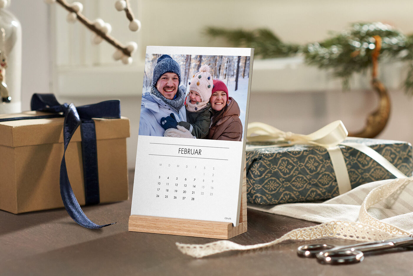 CEWE Fotokalender auf Holzaufsteller mit winterlichem Familienmotiv, umgeben von verpackten Geschenken.