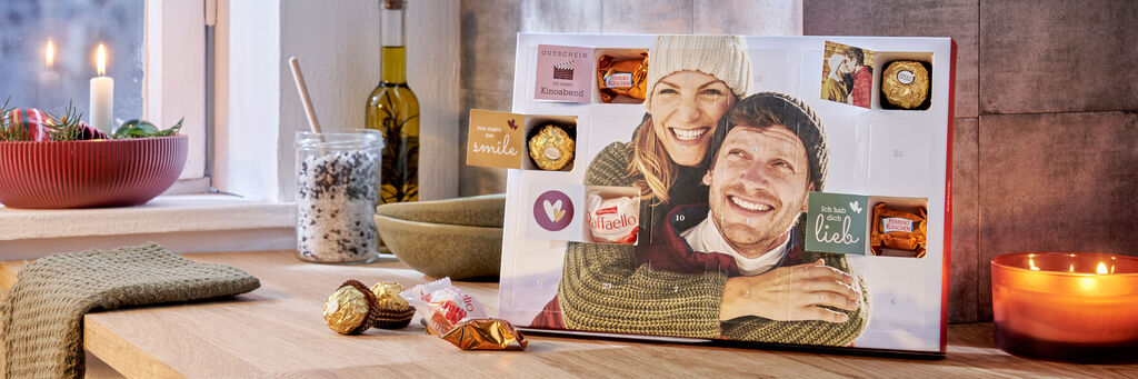 Ein Foto-Adventskalender steht auf einer Anrichte. Auf dem Cover sind eine Frau und ein Mann in winterlicher Kleidung zu sehen. Die Frau umarmt den Mann. Einige Türchen des Adventskalenders sind geöffnet und geben den Blick auf Ferrero-Pralinen frei. Auf den Innenseiten der Türen sind liebe Sprüche zu sehen. Neben dem Kalender stehen weihnachtliche Dekoration und eine Kerze.