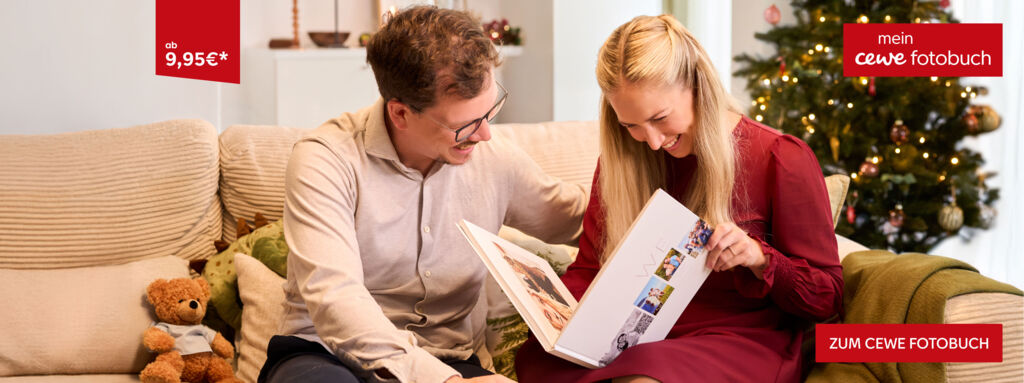 Zwei Personen blättern auf dem Sofa in einem CEWE FOTOBUCH. Dahinter ist ein Christbaum zu sehen.