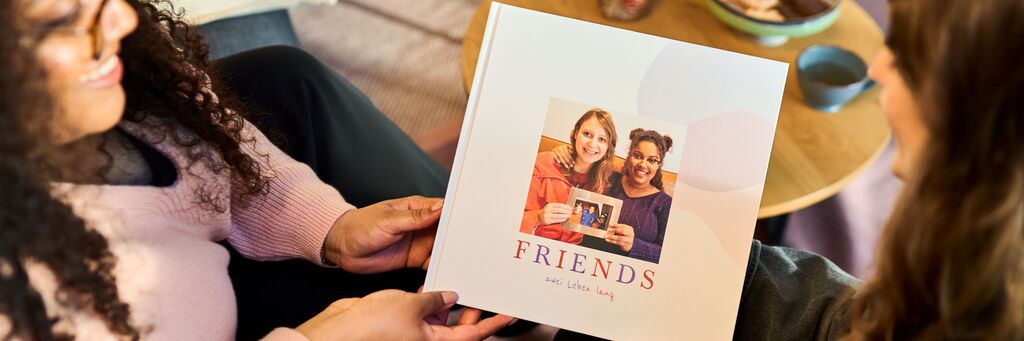 Die Aufnahme von schräg oben zeigt zwei junge Frauen im Halbprofil. Eine überreicht der anderen ein CEWE FOTOBUCH mit einem Foto der beiden sowie der Aufschrift "Friends". Im Hintergrund ist ein Wohnbereich mit einem kleinen Kaffeetisch zu sehen, auf dem Tee und Gebäck erkennbar sind.