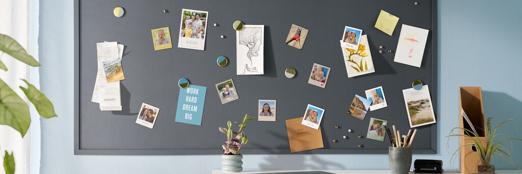 An einer Wand befindet sich eine graue Tafel, an der mehrere Fotomagnete im Retro-Stil sowie Postkarten und Fotos befestigt sind. Die Motive auf den Fotos und Karten zeigen Landschaften, lachende Menschen oder auch Schriftzüge. Vor der Tafel ist ein Schreibtisch zu erkennen.